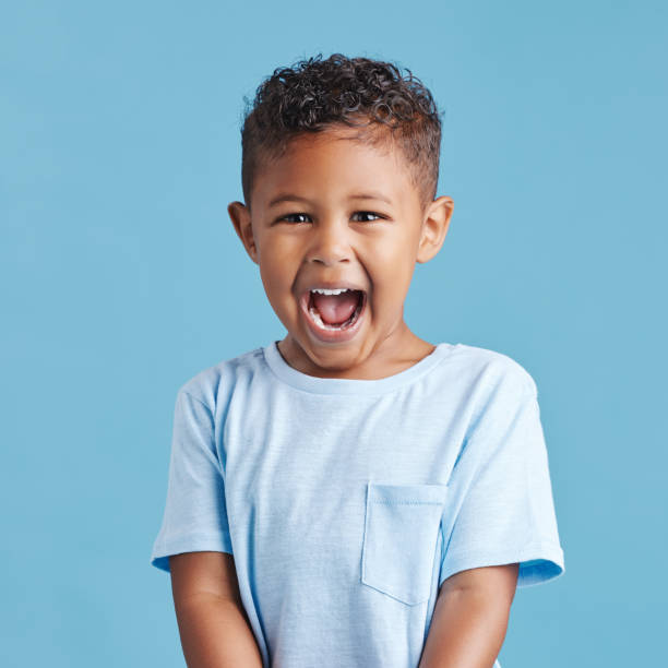 feche acima de menino animado olhando feliz e gritando enquanto está de pé contra um fundo de estúdio azul. pré-escolar bonito vestindo roupas casuais - child little boys isolated standing - fotografias e filmes do acervo