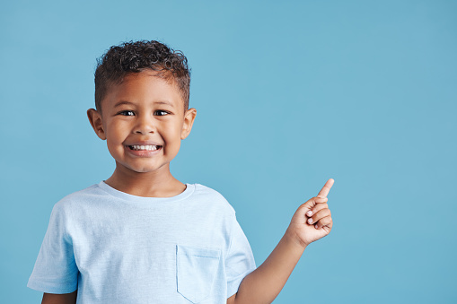 Portrait of happy smiling little boy pointing his finger at copy space to the right against blue studio background. Cheerful hispanic kid with healthy dental smile in casual clothes