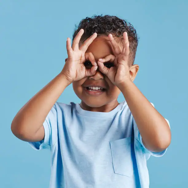 Photo of Portrait of inquisitive nosy little boy looking through fingers shaped like binoculars against a blue studio background. Curious child exploring