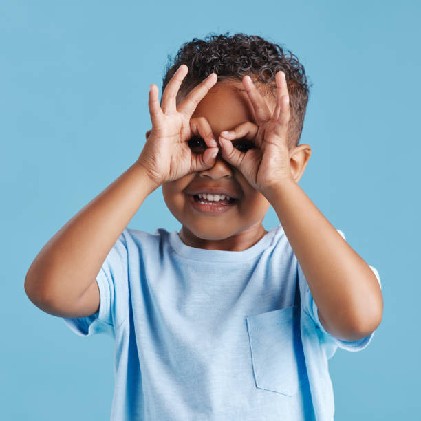 portret dociekliwego wścibskiego chłopca patrzącego przez palce w kształcie lornetki na niebieskim tle studyjnym. ciekawskie dziecko odkrywające - little boys discovery binoculars child zdjęcia i obrazy z banku zdjęć