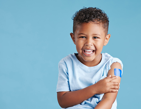 Happy vaccinated boy kid showing arm with adhesive bandage after vaccine injection standing against a blue studio background. Advertising vaccination against coronavirus. Child immunisation