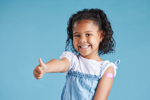 Happy vaccinated kid showing thumbs with plaster on arm after vaccine injection standing against a blue studio background. Advertising vaccination against coronavirus. Child immunisation