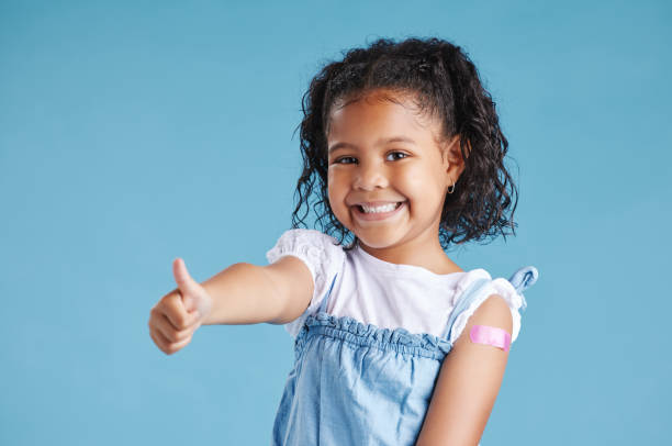 Happy vaccinated kid showing thumbs with plaster on arm after vaccine injection standing against a blue studio background. Advertising vaccination against coronavirus. Child immunisation Happy vaccinated kid showing thumbs with plaster on arm after vaccine injection standing against a blue studio background. Advertising vaccination against coronavirus. Child immunisation 4 year old girl stock pictures, royalty-free photos & images