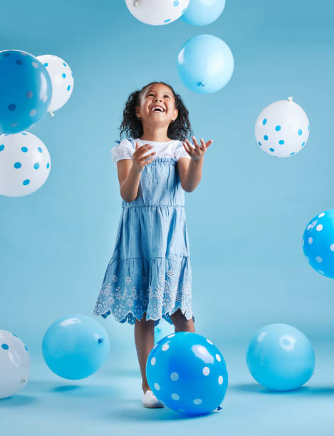 entzückendes kleines mädchen, das nach oben schaut, spielt und spaß mit blauen und weißen luftballons hat, um ihren geburtstag vor einem blauen studiohintergrund zu feiern - child balloon happiness cheerful stock-fotos und bilder