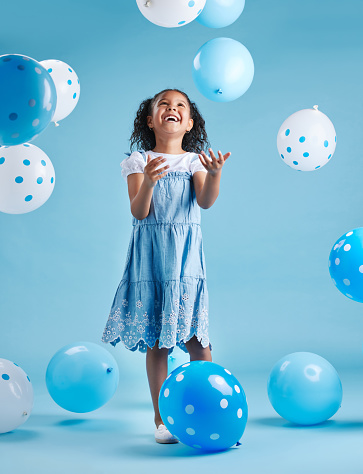 Adorable little girl looking up playing and having fun with blue and white balloons in celebration of her birthday against a blue studio background