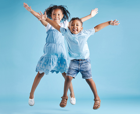 Full length portrait of cute girl and boy jumping with their arms outstretched and having fun on blue studio background. Happy little brother and sister siblings smiling and playing together