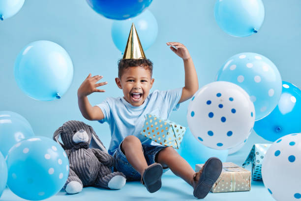 aufgeregter kleiner junge, der seinen geburtstag mit luftballons und geschenken auf blauem studiohintergrund feiert. süßes kind, das einen partyhut trägt und mit geschenken und einem ausgestopften hasen sitzt - child lifestyles isolated blue stock-fotos und bilder