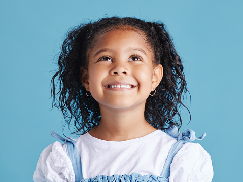 Close up head shot with smiling little brown haired girl looking up. Happy kid with good healthy teeth for dental on blue background