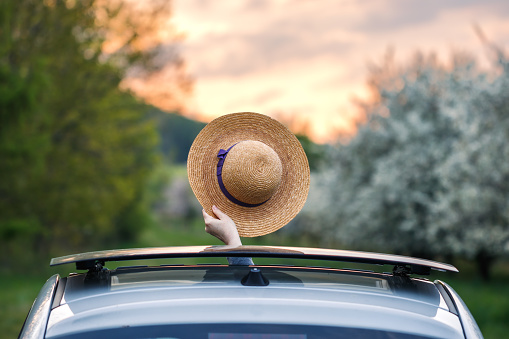 Woman waving with straw hat from car sun roof window during sunset. Road trip to adventure