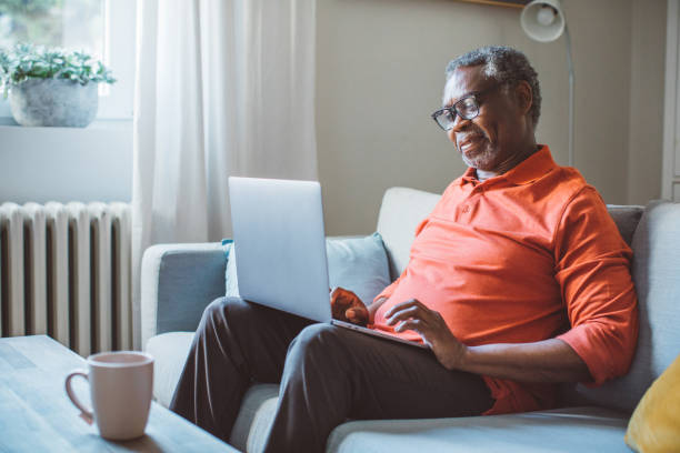Mature man using laptop Mature man at home, sitting on sofa and using laptop. black man laptop stock pictures, royalty-free photos & images