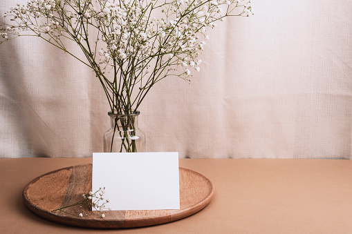 Blank card and gypsophila flowers in glass vase on wooden tray. Mockup.