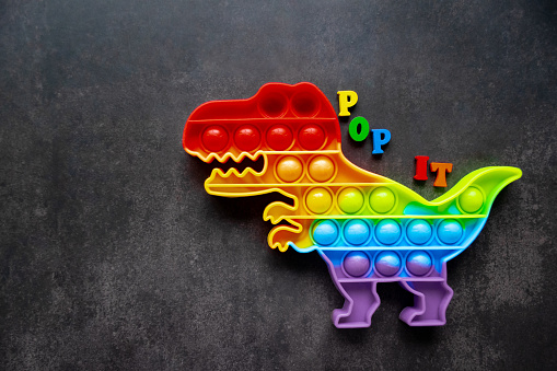 Toy pop it dinosaur rainbow colors on a black background with multicolored letters and the inscription-Pop it on the back.