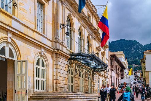 Bogota, Colombia - January 27, 2017: Looking up one of the narrow streets in La Candelaria in the Andean capital city of Bogota, in the South American country of Colombia. To the left is the Opera, Teatro Cristóbal Colón, and in the far background, the Andes Mountains. Some local Colombian people go about their daily chores. Photo shot in the afternoon sunlight on an overcast day; horizontal format. Copy space.