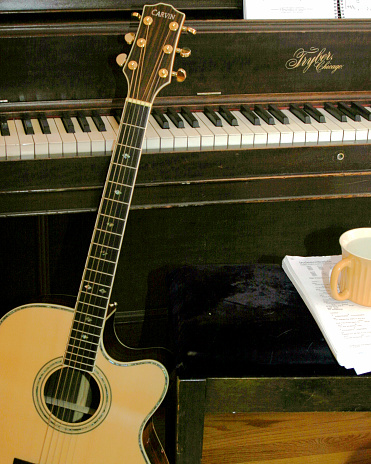 acoustic guitar leaning up agaisnt an upright grand piano, next to piano bench with sheet music and a mug of coffee on it.