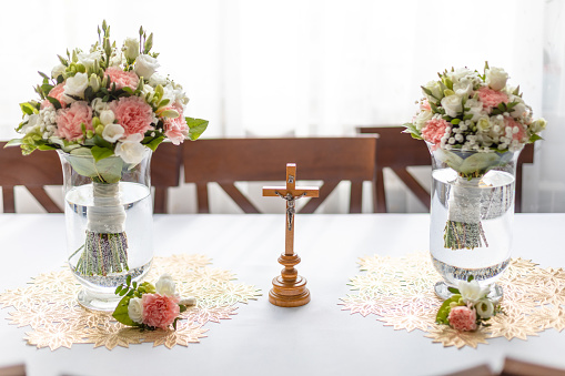 Soft bridal bouquets of fresh flowers in glass vases and church cross on celebratory table in restaurant. Elegant attribute and tradition. Beautiful and romantic wedding, luxurious celebration.