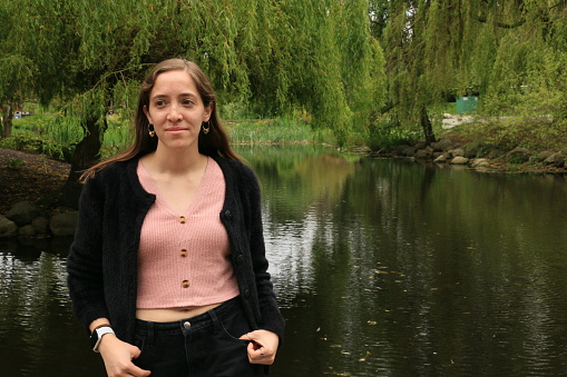 A Mexican woman near a pond. She is wearing a pink top, black sweater and pants.