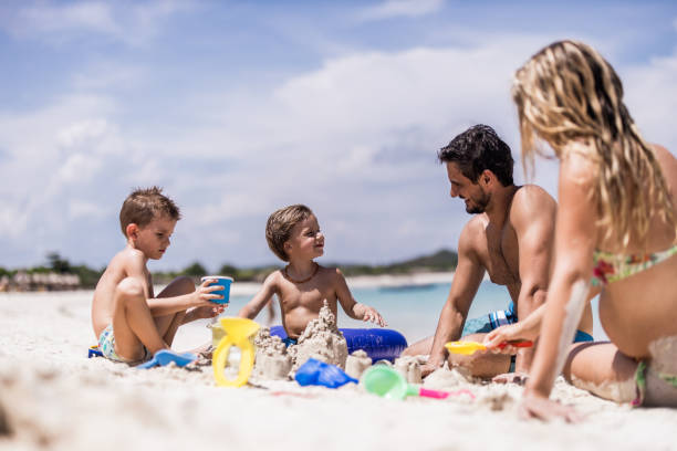 Young happy family playing with toys in sand on the beach. Happy parents and their small sons relaxing in sand on the beach and playing with toys. Copy space. family beach vacations travel stock pictures, royalty-free photos & images