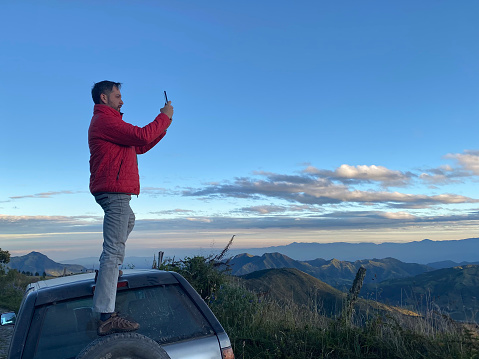 Young handsome latin male backpacker Photographer; hiking, enjoying and taking pictures using his smartphone with 5G technology to the Sierra's Highlands, in front of Pichincha, Cayambe and Cotopaxi's Volcanoes with a blue sky in a midday near to Quito, Pichincha's Province, Ecuador, Latin America. He's Capturing a beautiful view  from the top of a mountains.