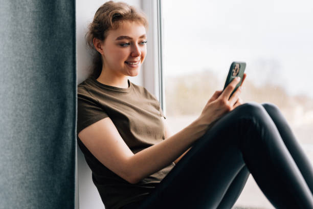 istock Happy young woman holds phone while sits on windowsill at home. 1399640917