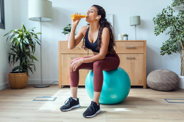 giovane donna sportiva che beve succo disintossicante mentre fa esercizio con la palla a casa. - yoga ball foto e immagini stock