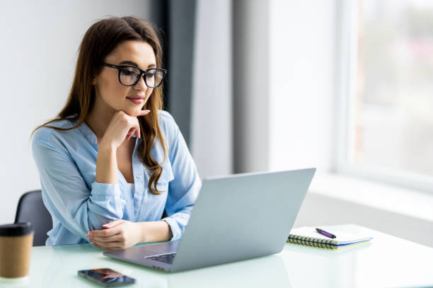 istock Young woman with laptop computer sitting at table over office window background 1399647272