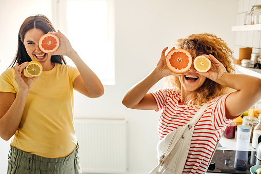Two beautiful girls are covering eyes with slices of orange and smiling while standing in their kitchen at home