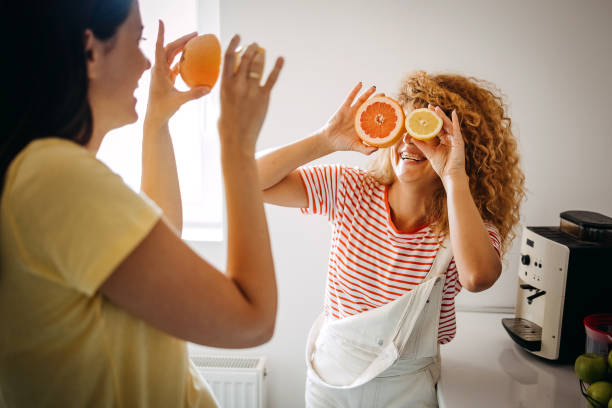 Two beautiful girls are covering eyes with slices of orange and smiling while standing in their kitchen at home