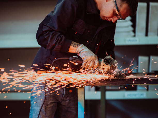 istock Heavy Industry Engineering Factory Interior with Industrial Worker Using Angle Grinder and Cutting a Metal Tube. Contractor in Safety Uniform and Hard Hat Manufacturing Metal Structures. 1399657755