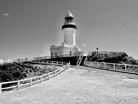 Horizontal black and white landscape photo of the iconic 22m high Cape Byron lighthouse built in 1901 on the rugged Cape Byron Headland, Australia’s most easterly point. Byron Bay, north coast NSW. The walking track, coastal scrub forest, a white painted wooden fence and a mown lawn area are in the foreground.