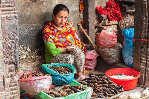 Kathmandu, Nepal - October 27, 2021: Hindu woman sells dried fish in the streets of Kathmandu. Woman dressed in colored clothes is waiting for customers. Between the old decaying houses of Kathmandu