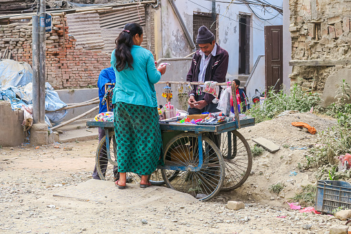 Kathmandu, Nepal - October 27, 2021:  Street trader sells his penny goods in the ruins of the earthquake stricken city. Woman checks the goods of the rickshaw like stall. Street scene in the capitol