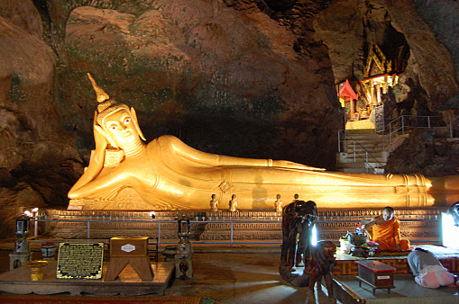 Woman praying in front of Cross-legged monk by the reclining Buddha at Cave temple (Wat Tham Suwan Khuha), in Phang-Nga province, Thailand