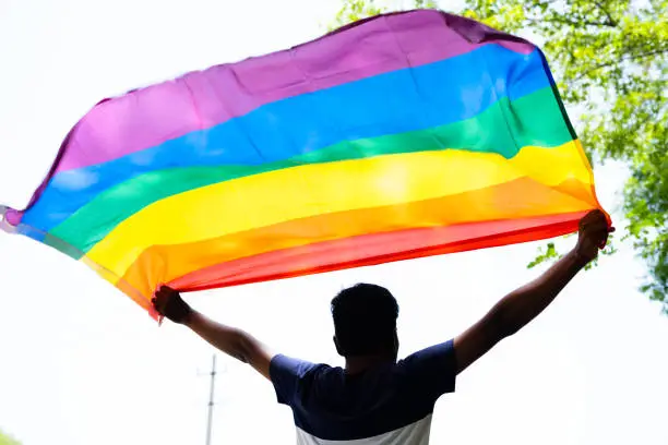 back view shot of young man proudly holding waving lgbtq flag - concept of pride month celebration, support and movement