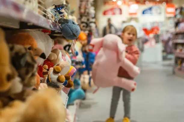 Little caucasian girl choosing a new stuffed animal toy in the big baby store. Big shelfs full of toys