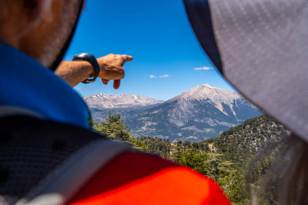 Close-up shot of two people in backpacks hats side by side, man with wristwatch with their backs turned, showing mountains with snowy peaks admiring the view.Small clouds in places