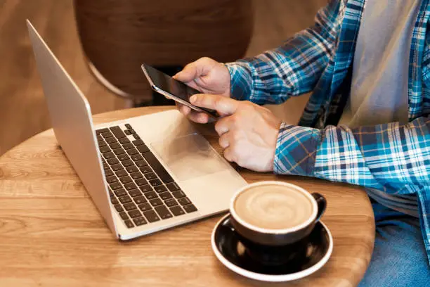 Photo of Casual man freelancer working online with macbook and mobile phone and drinking morning coffee in cafe