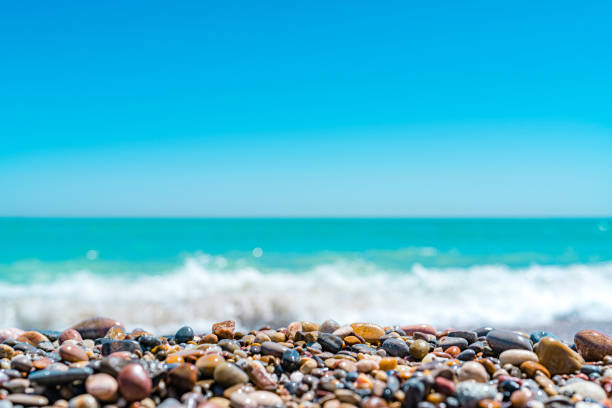 Summer abstract backgrounds: blurred tropical turquoise beach with white waves at background with focused sea pebbles at the bottom of the frame.