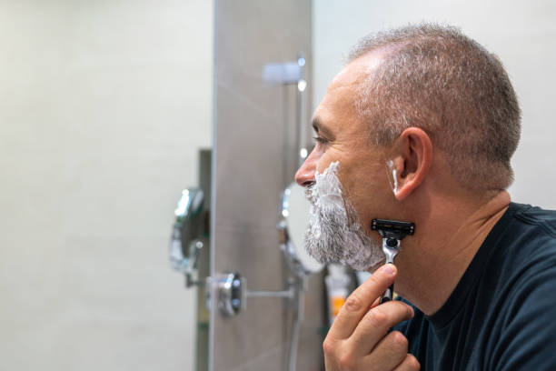 istock Handsome white-haired beared man shaving off his beard looking in a mirror. Concept of changing style 1399704578