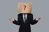Young businessman covering his face using a craft paper bag with red drawn question mark, like a mask, for hiding his identity. Man isolated on gray wall background.