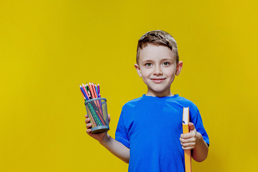 Smiling happy schooler in blue t-shirt holding multicolored pencils and book on yellow background.