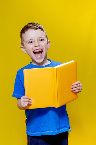 Positive preschool boy in an blueT-shirt holding an open yellow copybook on yellow background.