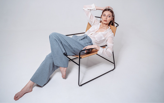 A beautiful young woman full height  in pants and a white shirt sits barefoot  in a chair against the background of the studio.