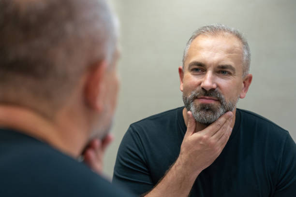 istock Middle-aged handsome smiling man looking in mirror in bathroom touching his beard thinking of cutting it off 1399708177