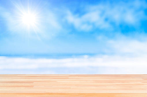 Summer abstract backgrounds. Empty wooden table at the bottom of the frame with defocused blue sky with bright sun and beach at background. Diminishing perspective on table. Focus on table. Copy space, ideal for product montage.