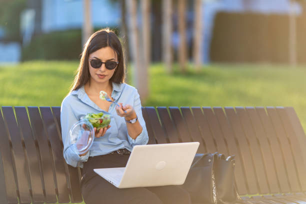 istock Business woman is sitting on bench outdoors and using lap top for working out office and eating salad 1399714253