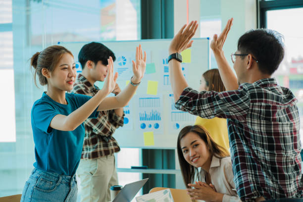 istock Young Asian business groups are feeling happy and successful. Laughing hands up celebrating achievements on job goals deal with laptop on desk and background finance graph in office. 1399727607