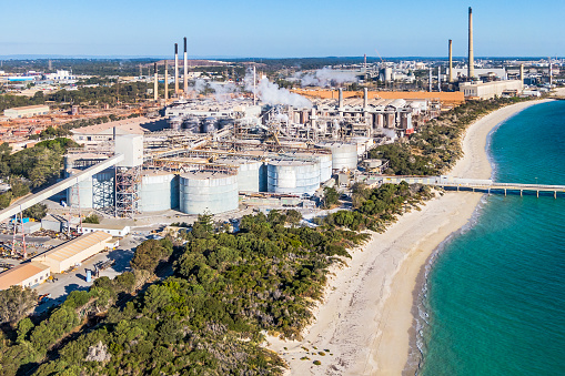 Aerial view alumina refinery in close proximity to natural coastal scrub, clean sandy beach and clear turquoise Indian Ocean sea. Large manufacturing complex with jetty jutting out to sea.