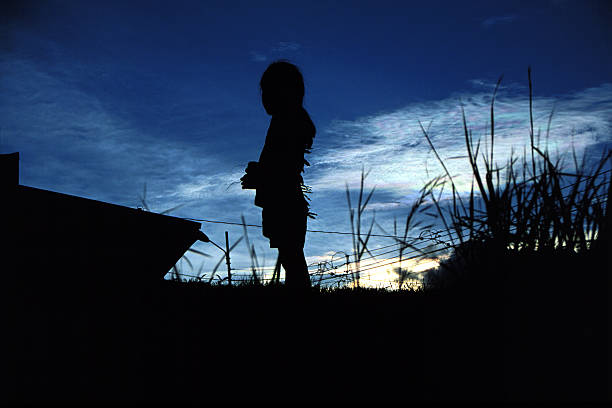 Child silhouetted against a bright blue sky and some low clouds.  Nearby are silhouettes of tall grass and a small building. Shot in the barrios (slums) of Costa Rica.