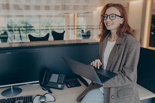 Beautiful woman office worker in casual clothes wearing glasses, sitting on desk at her workplace in office, using laptop computer and smiling, enjoying nice working day. Happy business woman at work