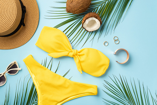 Summer concept. Top view photo of sunhat coconuts gold rings bracelet stylish sunglasses yellow bikini and palm leaves on isolated pastel blue background